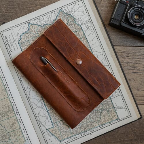 Exterior cover of the brown bison leather Journal Cover from Duvall Leather that features an exterior pen pocket stamped with "Duvall", laying on top of an atlas on a wood surface with a camera next to it.