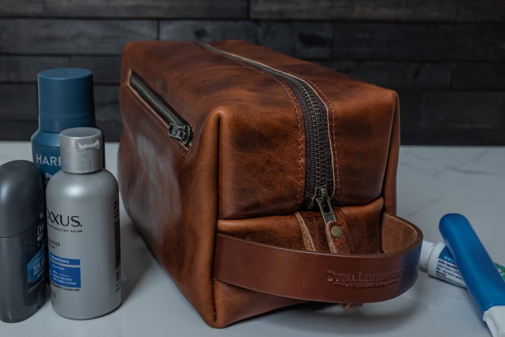 A brown bison leather toiletry bag or Dopp kit sitting on a white marble bathroom counter along with a few toiletries.