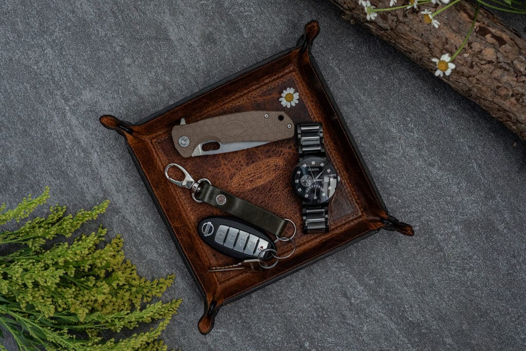 Brown bison leather valet tray on a grey granite counter top with spring flowers filled with a dress watch, pocket knife, and a set of keys.