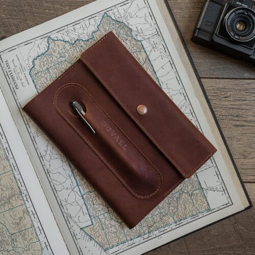 Exterior cover of the red brown leather Journal Cover from Duvall Leather that features an exterior pen pocket stamped with "Duvall", laying on top of an atlas on a wood surface with a camera next to it.