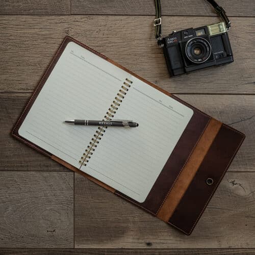 Red Brown Leather Journal Cover with a notebook inside laid out on a wood surface with a gray Duvall Leather pen laying on top of the notebook.