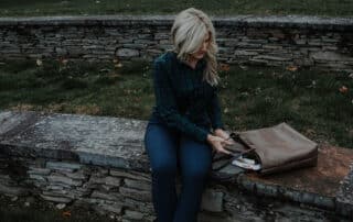 A woman taking a wallet out of a taupe large leather tote bag while sitting outside on a stone bench