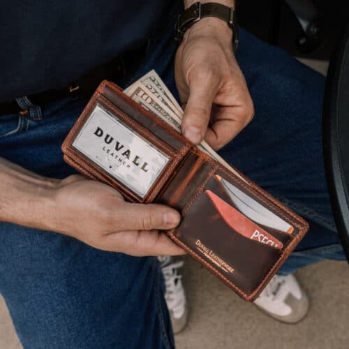 Man holding open a brown leather bifold wallet open to show the inside card holders and ID window while also pulling some cash out of the wallet.