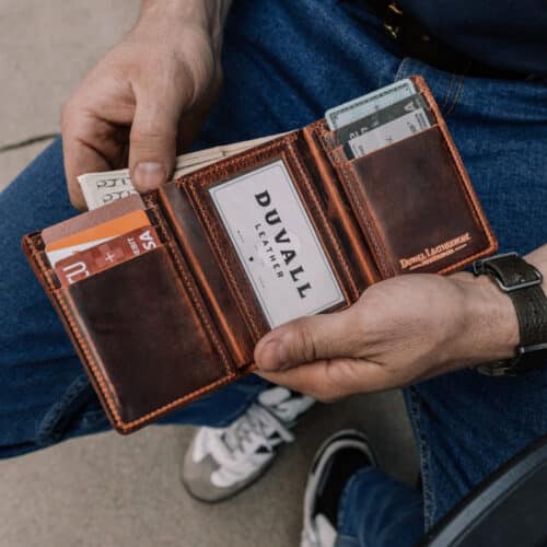 Man holding open a brown leather trifold wallet open to show the inside card holders and ID window while also pulling some cash out of the wallet.