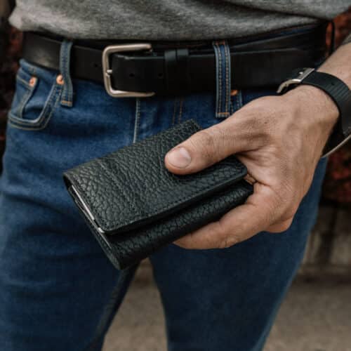 Man holding a closed black bison leather trifold wallet in his hand.