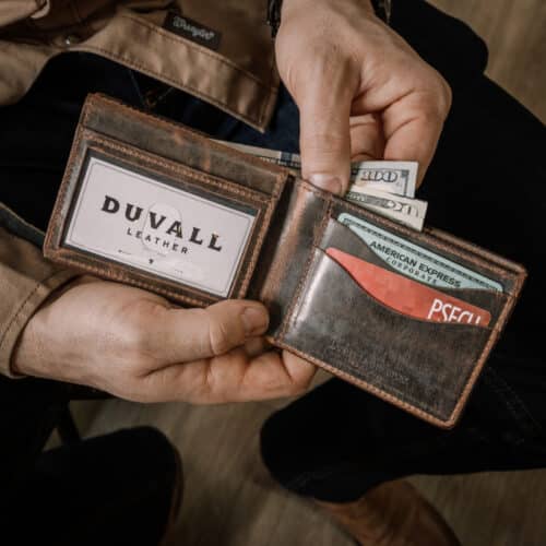 A man's hands are holding open a brown leather deluxe bifold wallet with a vintage finish showing the cards, cash, and ID window inside.