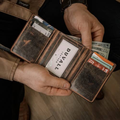 A man's hands are holding open a brown leather trifold wallet with a vintage finish showing the cards, cash, and ID window inside.