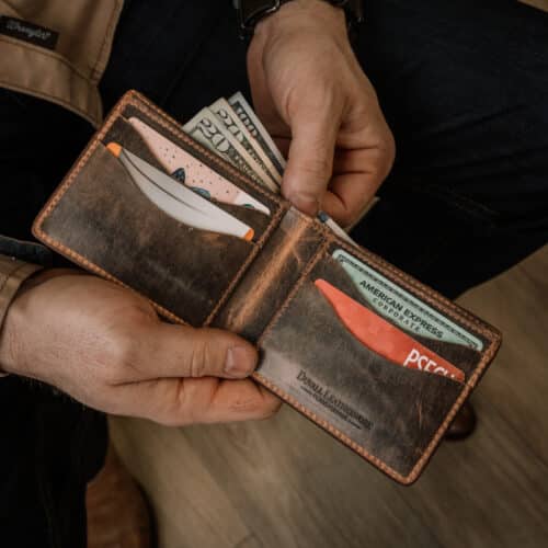 A closeup of a man holding open a brown leather bifold wallet with a vintage finish showing that this wallet has ample room for both cards and cash inside.