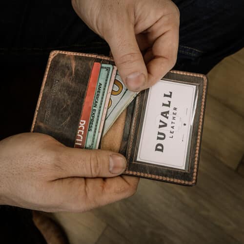 A man's hands are holding open a brown leather credit card ID window wallet with a vintage finish showing the cards, cash, and ID window inside.