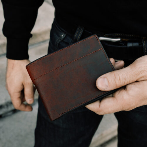 A close up image of a man holding a closed, dark reddish-brown leather bifold wallet in his hand to showcase the exterior of the wallet.