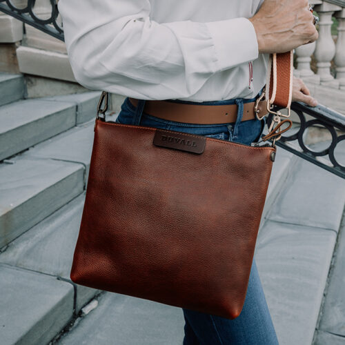 Close up of a woman dressed in a white dress shirt and blue jeans carrying a red brown leather crossbody bag.