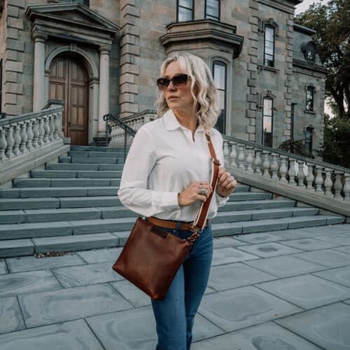 Woman with sunglasses wearing a white button down shirt and blue jeans is sporting a red brown leather crossbody bag.