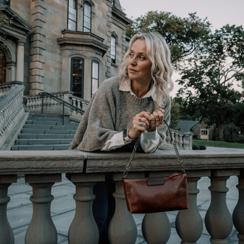 Woman wearing a light brown poncho sweater holding a small red brown leather handbag that has a short shoulder strap made of leather and elegant metal chains.