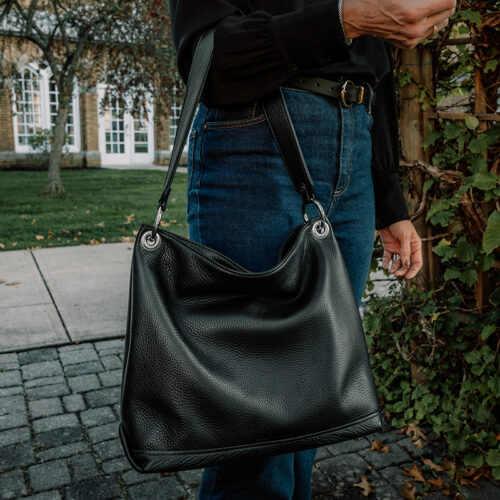 A close-up photo of a black leather slouch bag held by a woman wearing a black blouse and blue jeans.