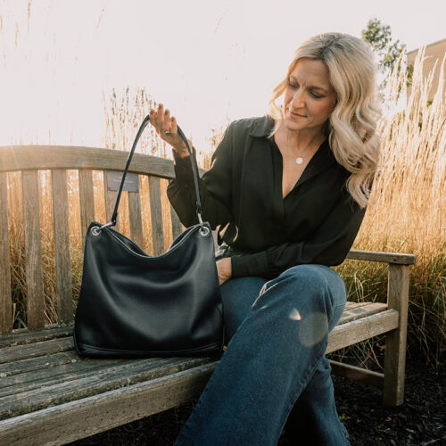 A photo of a woman holding the handle of a black leather slouch bag while sitting on a bench next to the bag. The woman is wearing a black blouse and blue jeans.