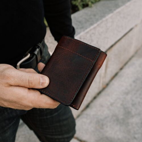 Close up of a man holding a closed, dark reddish-brown leather trifold leather in one hand.