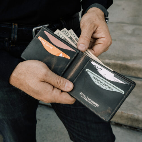 A close up of a man holding an open bifold wallet showcasing the dark grey leather interior along with the card sleeves and cash pocket.