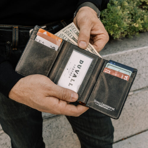 A close up image of a man holding a leather trifold wallet open to the dark grey cowhide leather inside showcasing the ID window, card slots, and cash.
